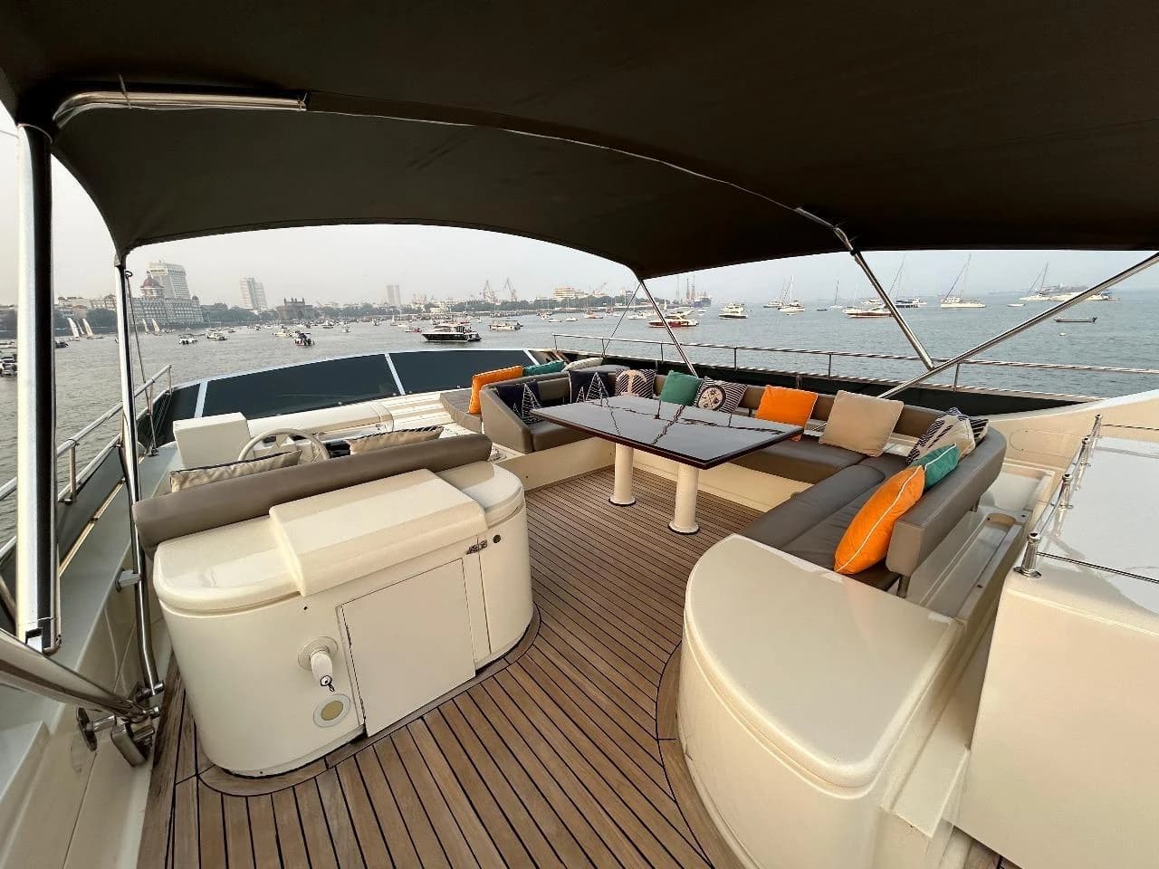 Flybridge lounge seating under the dark bimini, with the Mumbai skyline and harbour boats behind