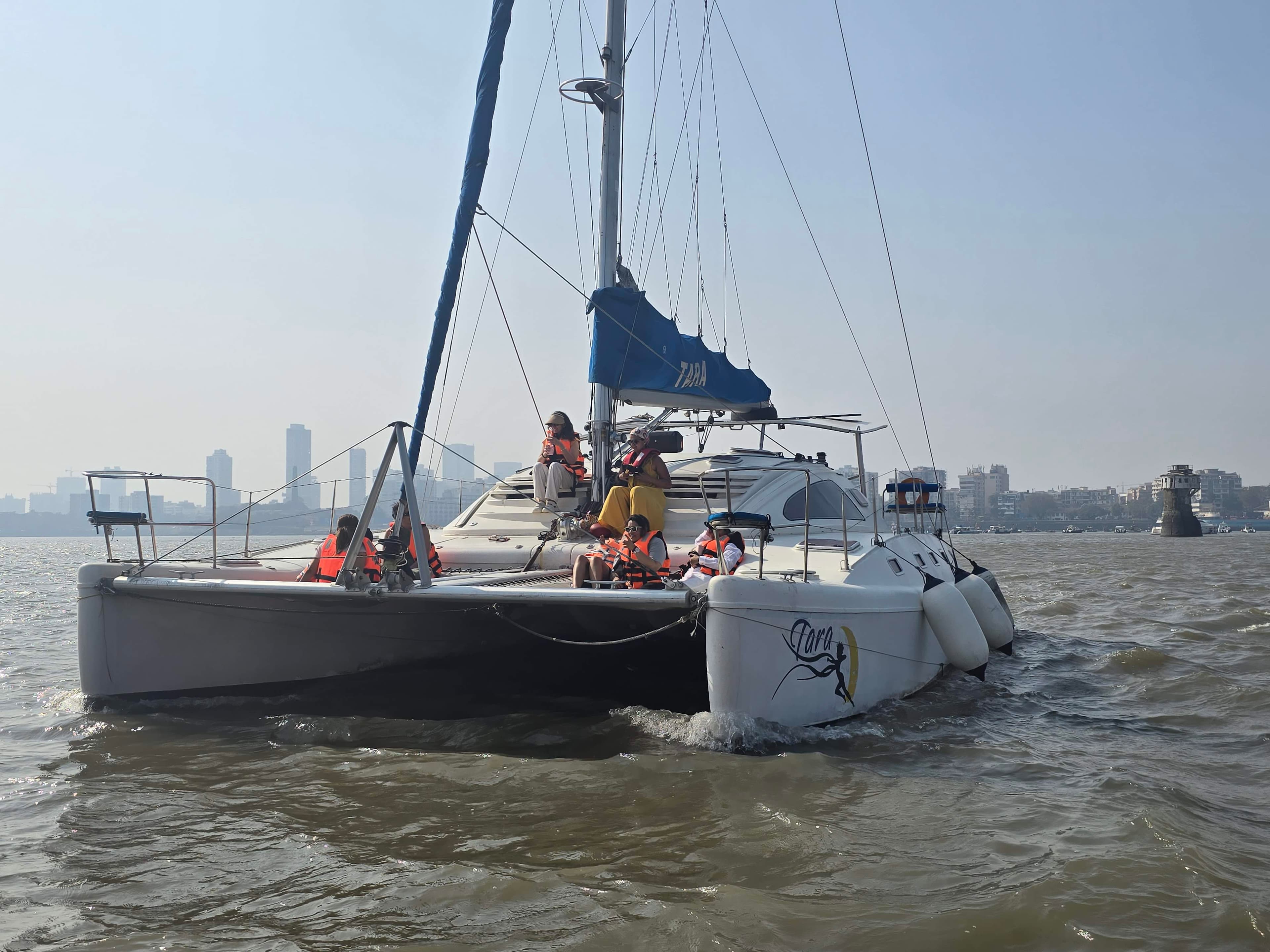 Guests in life jackets aboard Tara under sail with the Mumbai skyline behind