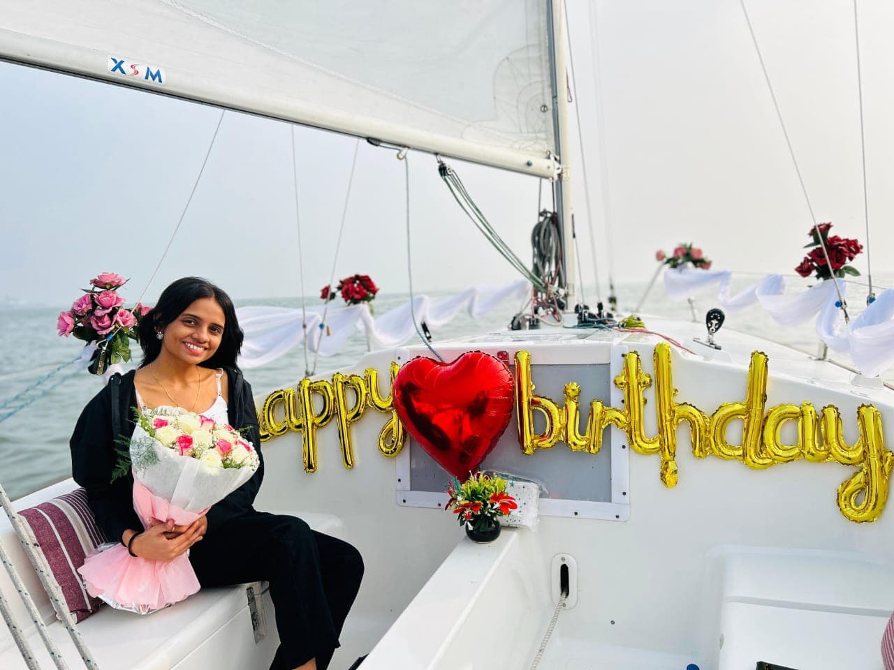 A guest seated at the bow holding a rose bouquet beside a gold 'Happy Birthday' balloon display and a red heart balloon