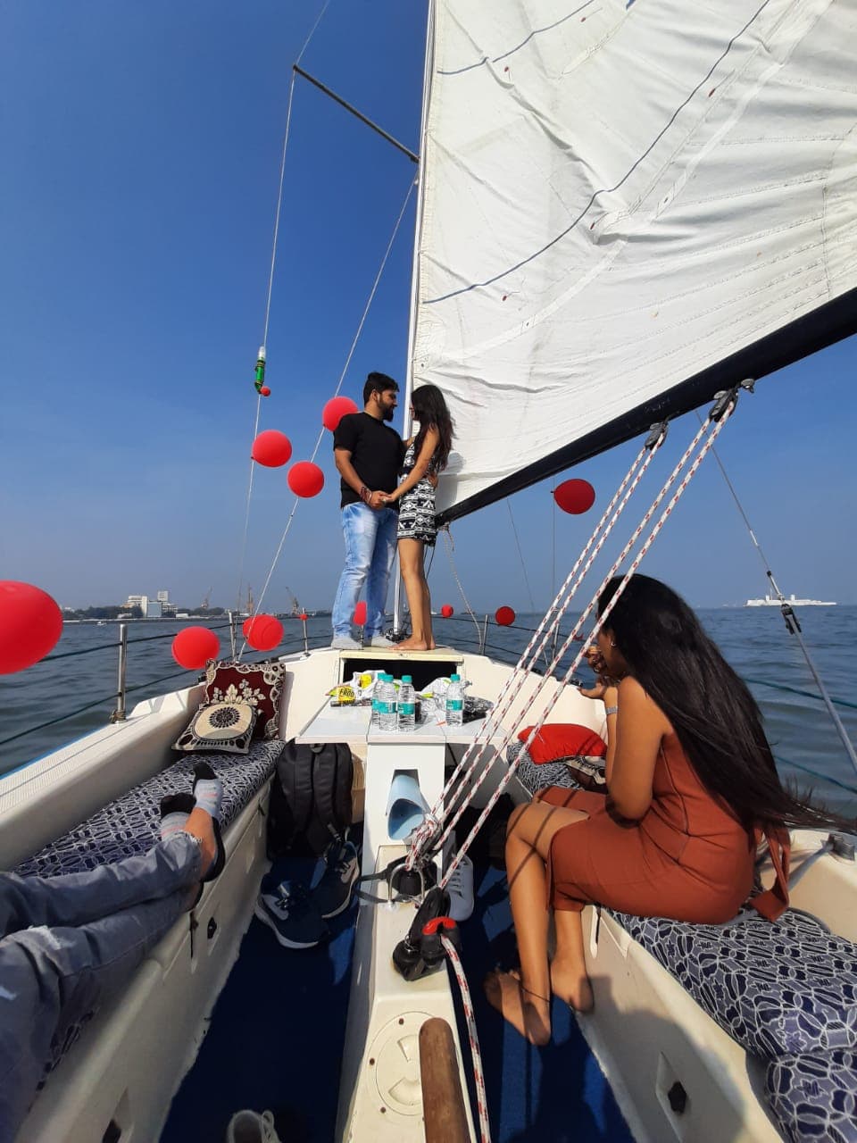 A couple standing at the bow under sail, decorated with red balloons, Mumbai skyline in the distance