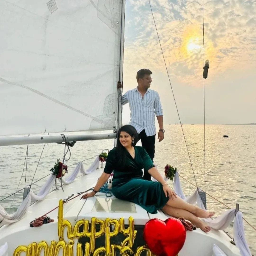 A couple posing on deck at golden hour with 'Happy Anniversary' balloons and fresh flowers along the rail
