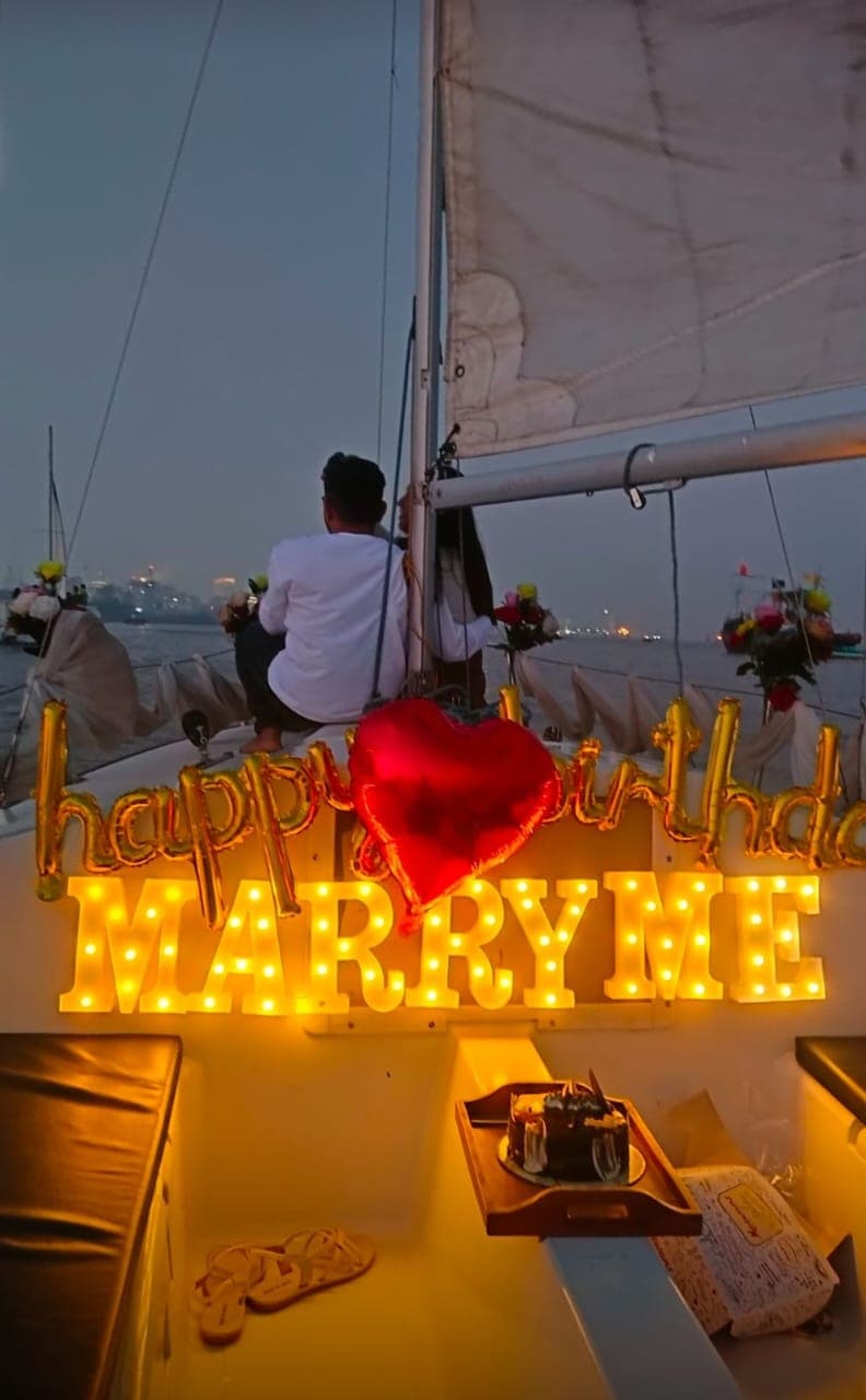 An illuminated 'Happy Birthday, Marry Me' balloon display at dusk with a cake and fairy lights on deck