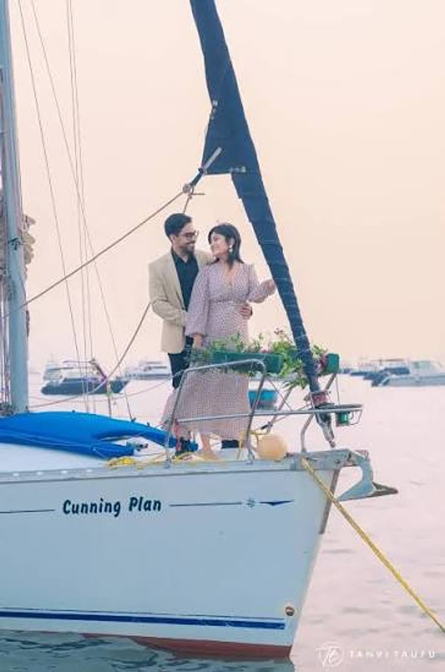 A couple standing quietly at the mast of a sailboat at dusk, the Mumbai skyline behind