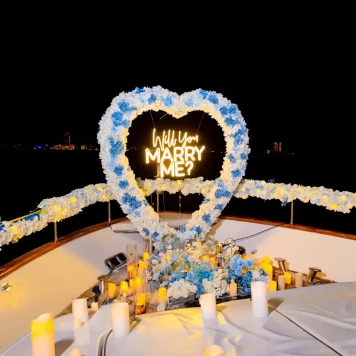 A heart-shaped floral arch with an illuminated Will You Marry Me sign and candles on a yacht bow at night