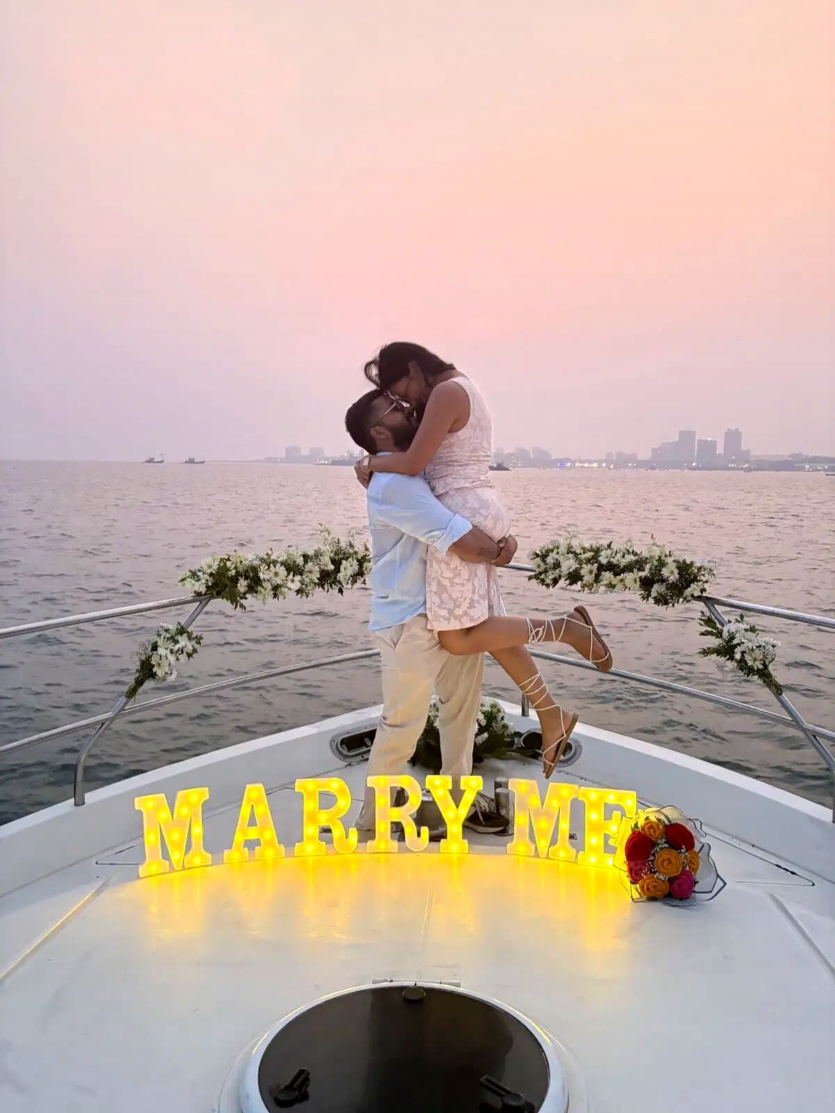 A couple embracing beside a lit Marry Me sign on a yacht bow at sunset, the Mumbai skyline behind